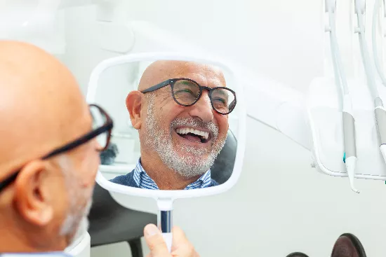 older man smiling in a mirror, showing his new dental implants