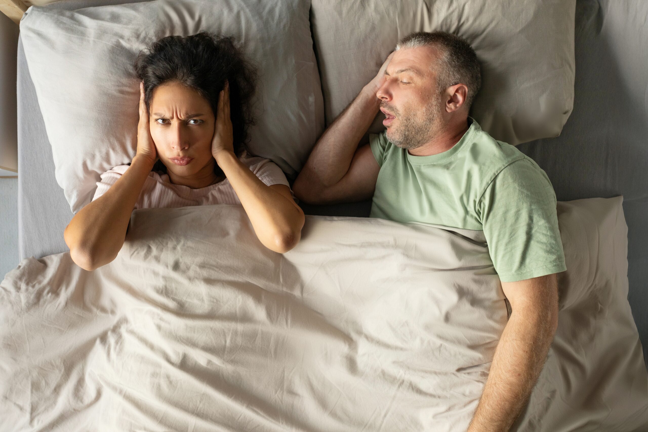 Annoyed woman closing ears with hands suffering from loud snore of her sleeping husband lying nearby